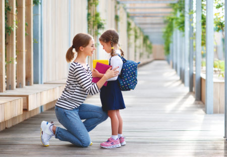 A little girl with pigtails standing on a path at school, wearing a backpack and holding books. Her mother is kneeling down facing her, holding her shoulders and smiling.