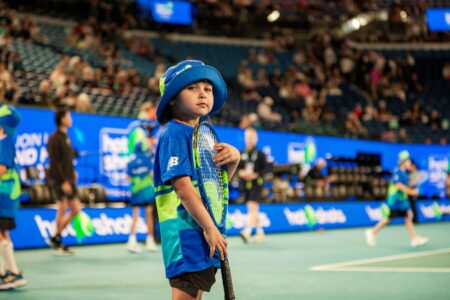 A child on a tennis court holds a racket and looks at the camera. Behind them, the stadium is starting crowd is starting to fill up. 