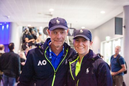 A man and a woman look at the camera smiling. They're both wearing matching Australian Open branded uniforms and caps.