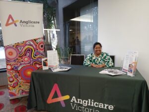 Jaya Mathew is smiling and sitting behind a desk with Anglicare Victoria branding at the AAFT conference