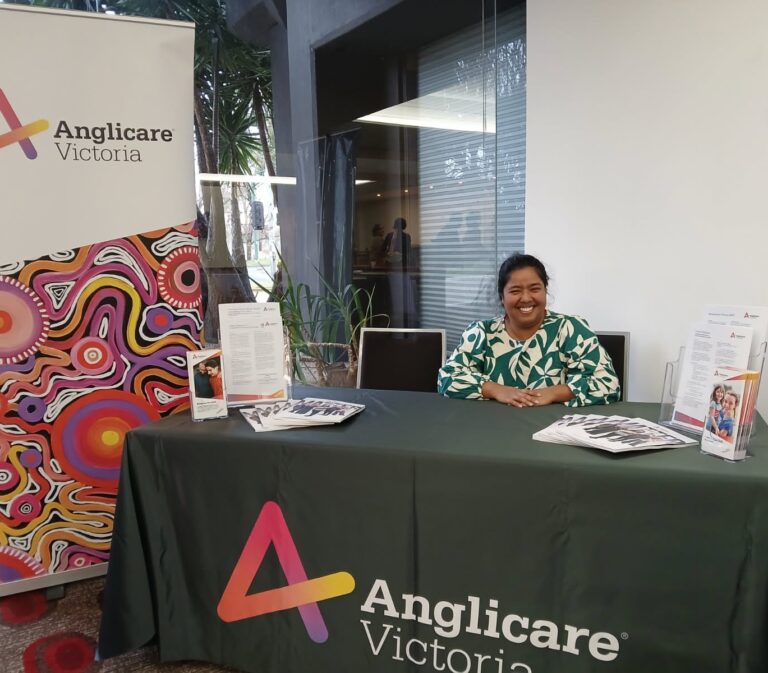 Jaya Mathew is smiling and sitting behind a desk with Anglicare Victoria branding at the AAFT conference