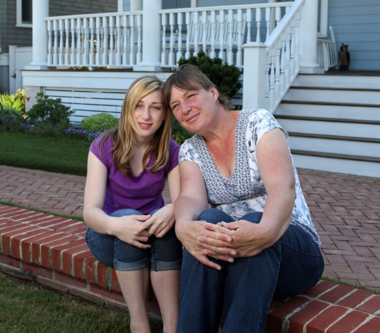 A teenage girl and a woman sitting together in front of a house