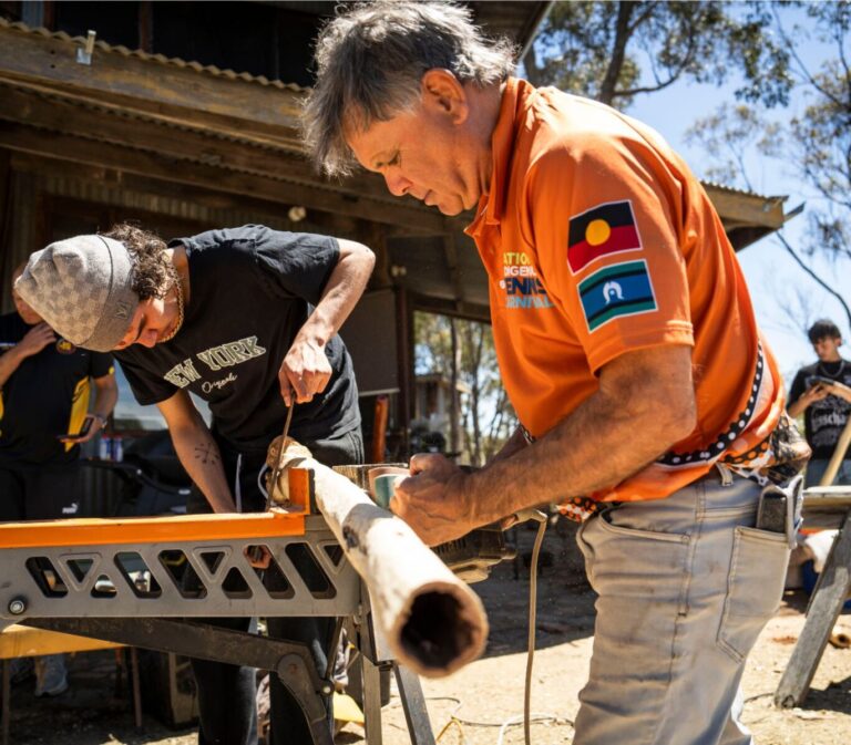 Elder Uncle Ron Murray sanding the bark from a didgeridoo with young person.