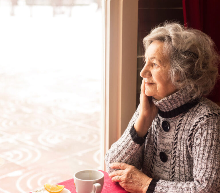 An older woman sits at a table with a cup of tea, looking out a window